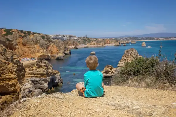 Kleinkind blickt auf die Küste Portugals. Felsen, türkisfarbenes Meer und eine Bucht mit Strand.