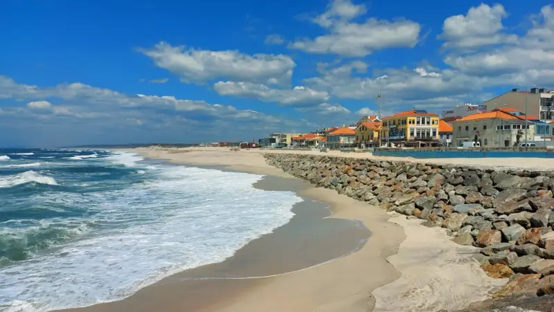 Sonne, Sand und Wellen an der Costa Verde, Portugal. Häuser mit roten Dächern säumen den Strand, geschützt von einer Steinmauer.